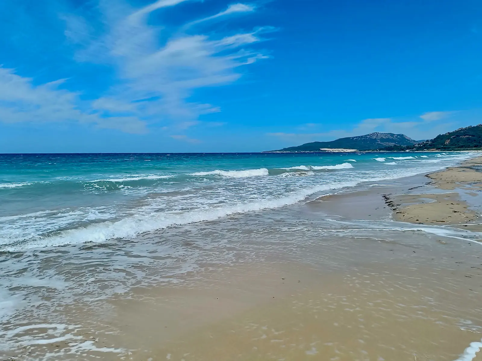 Sandstrand mit sanften Wellen und türkisem Meer unter blauem Himmel, im Hintergrund Berge